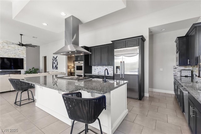 kitchen featuring a breakfast bar area, dark stone countertops, built in appliances, island exhaust hood, and decorative backsplash