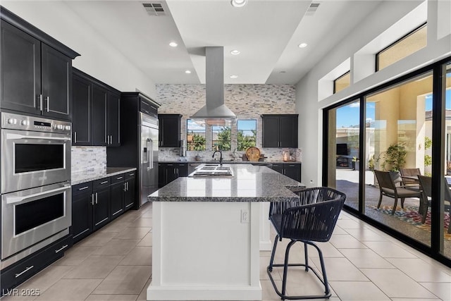 kitchen with appliances with stainless steel finishes, light stone countertops, a center island with sink, and plenty of natural light