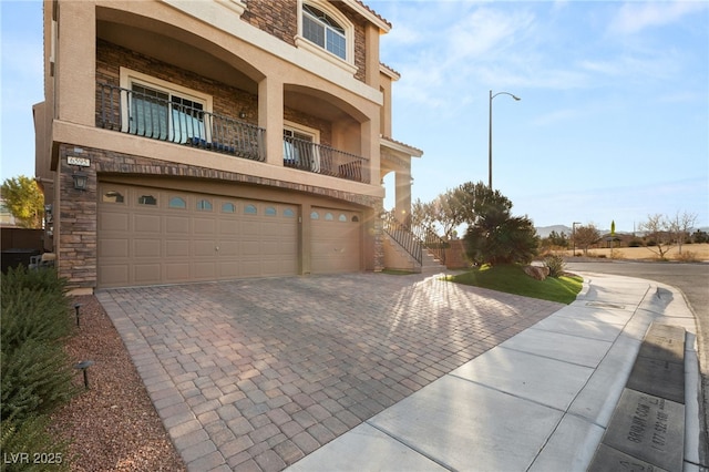 view of front of property featuring stucco siding, decorative driveway, stone siding, an attached garage, and a balcony