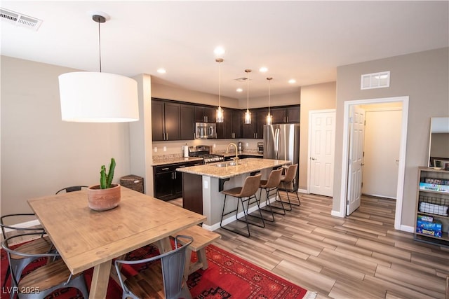 kitchen with sink, appliances with stainless steel finishes, an island with sink, decorative light fixtures, and light wood-type flooring