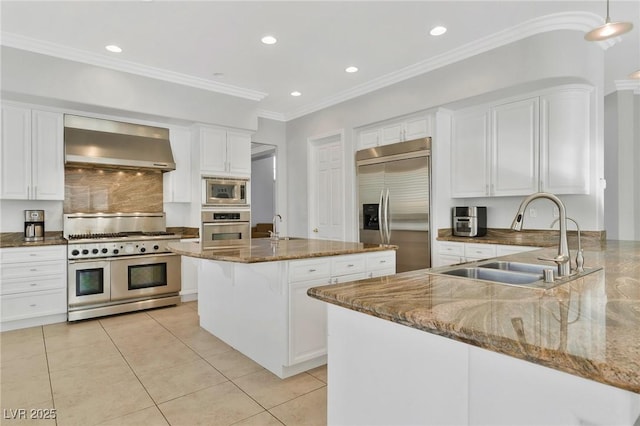 kitchen with wall chimney exhaust hood, sink, built in appliances, dark stone countertops, and an island with sink