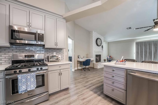 kitchen featuring tasteful backsplash, vaulted ceiling, a brick fireplace, light wood-type flooring, and stainless steel appliances