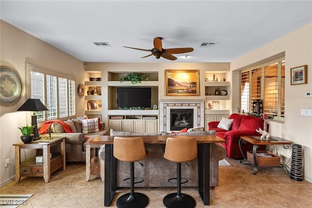 living room with built in shelves, ceiling fan, a stone fireplace, and light tile patterned floors