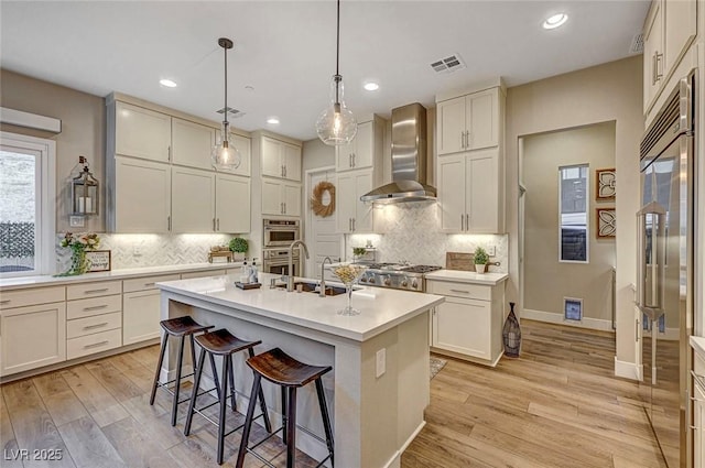 kitchen featuring pendant lighting, light hardwood / wood-style floors, an island with sink, high end refrigerator, and wall chimney exhaust hood