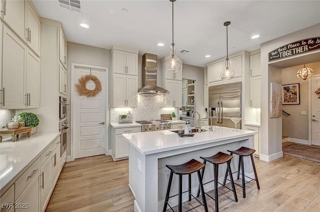 kitchen with pendant lighting, wall chimney range hood, appliances with stainless steel finishes, a kitchen island with sink, and light wood-type flooring