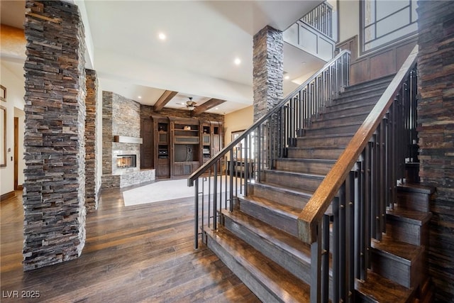 stairs featuring ornate columns, beam ceiling, ceiling fan, a stone fireplace, and hardwood / wood-style flooring