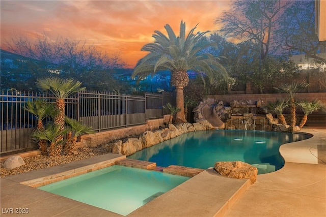 pool at dusk with a pool with connected hot tub, a water slide, and fence