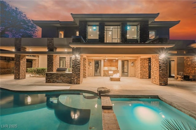 back of house at dusk with area for grilling, a pool with connected hot tub, stucco siding, stone siding, and a patio area