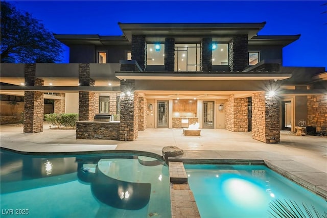 back of house at night featuring a pool with connected hot tub, stucco siding, stone siding, an outdoor kitchen, and a patio