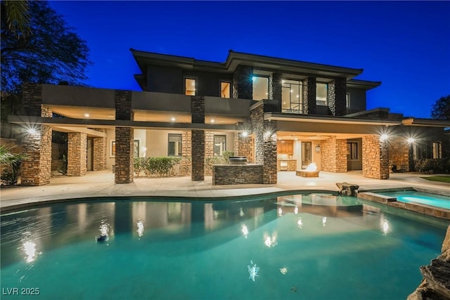 back of house at night with stucco siding, a pool with connected hot tub, stone siding, a patio, and a balcony