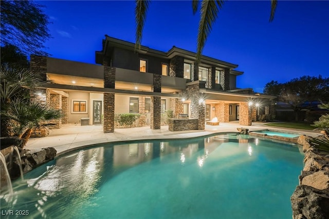 back of house at night featuring stone siding, stucco siding, a pool with connected hot tub, and a patio