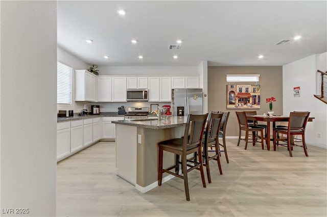 kitchen with white cabinetry, light wood-type flooring, dark stone countertops, an island with sink, and stainless steel appliances