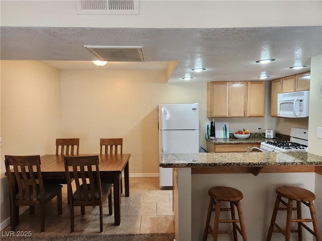 kitchen with stone countertops, a kitchen bar, white appliances, light brown cabinets, and a textured ceiling