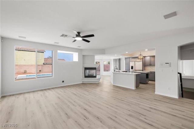 unfurnished living room featuring light wood-style flooring, a sink, a multi sided fireplace, baseboards, and a ceiling fan