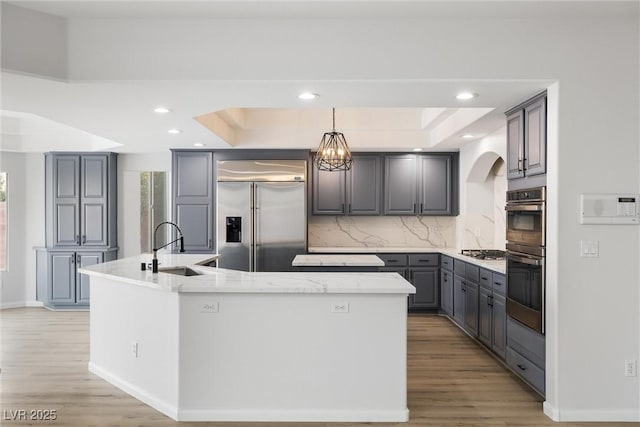 kitchen with a sink, appliances with stainless steel finishes, gray cabinets, an island with sink, and a raised ceiling