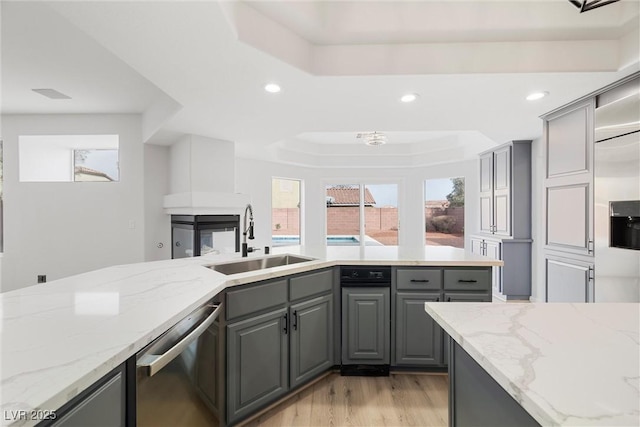 kitchen with a sink, stainless steel dishwasher, gray cabinets, light stone countertops, and a tray ceiling