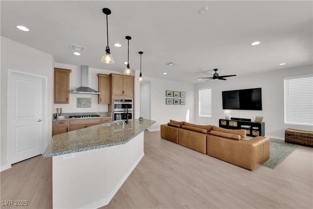 kitchen with wall chimney exhaust hood, sink, light stone counters, a center island with sink, and pendant lighting