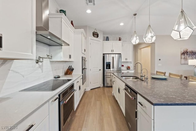 kitchen with appliances with stainless steel finishes, sink, white cabinets, a kitchen island with sink, and wall chimney range hood