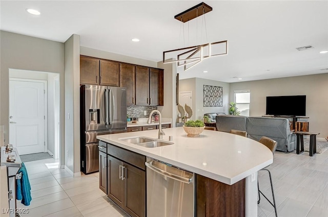 kitchen featuring sink, appliances with stainless steel finishes, hanging light fixtures, dark brown cabinetry, and a center island with sink