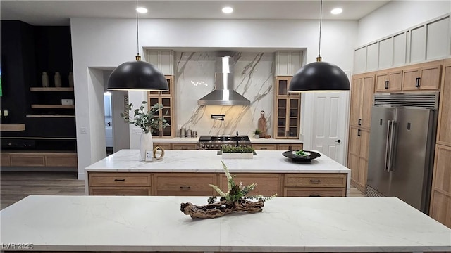 kitchen featuring wall chimney range hood, a kitchen island with sink, high quality fridge, light stone countertops, and decorative light fixtures