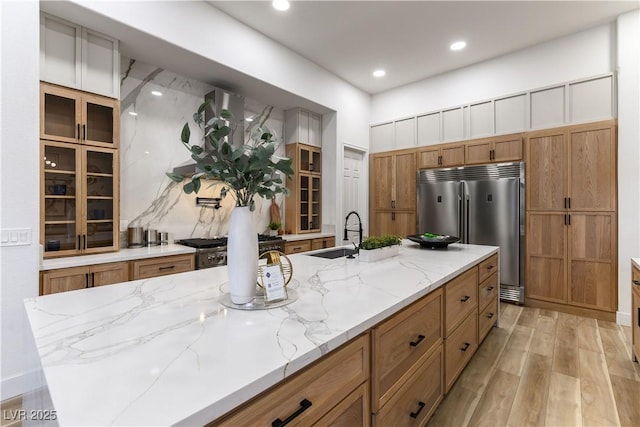 kitchen featuring sink, a large island with sink, stainless steel built in fridge, light stone counters, and light wood-type flooring