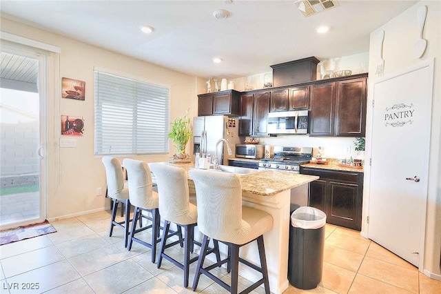 kitchen featuring a breakfast bar area, appliances with stainless steel finishes, dark brown cabinetry, a center island with sink, and light tile patterned flooring