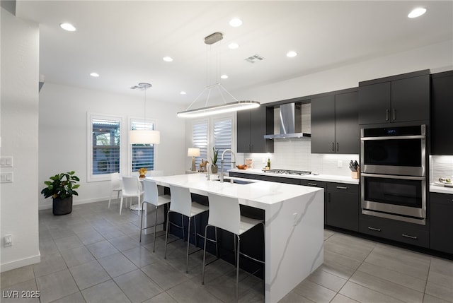 kitchen with sink, appliances with stainless steel finishes, an island with sink, decorative light fixtures, and wall chimney exhaust hood