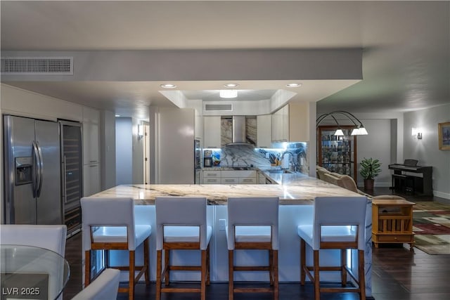 kitchen with a breakfast bar area, stainless steel fridge, kitchen peninsula, wall chimney range hood, and white cabinets