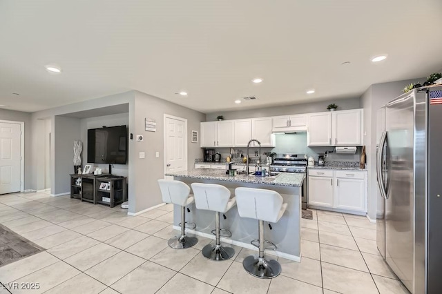 kitchen with appliances with stainless steel finishes, white cabinetry, a kitchen breakfast bar, light stone countertops, and a center island with sink