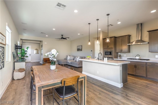 kitchen with hanging light fixtures, stainless steel microwave, gas stovetop, an island with sink, and wall chimney exhaust hood