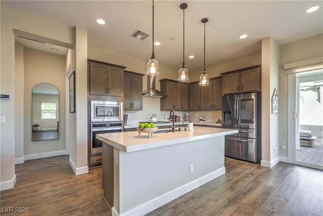 kitchen with decorative light fixtures, dark brown cabinets, appliances with stainless steel finishes, a kitchen island with sink, and wall chimney range hood