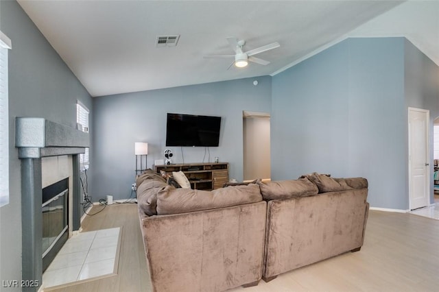 living room with ceiling fan, a tiled fireplace, vaulted ceiling, and light wood-type flooring