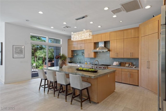 kitchen with hanging light fixtures, tasteful backsplash, an island with sink, wall chimney exhaust hood, and light brown cabinets