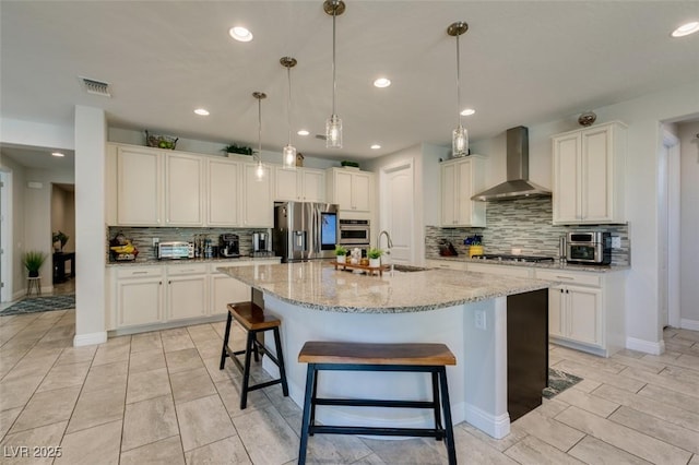 kitchen featuring wall chimney range hood, a center island with sink, hanging light fixtures, and appliances with stainless steel finishes