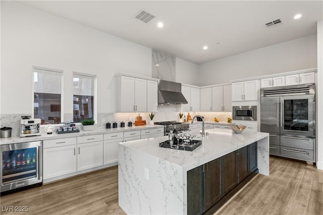 kitchen featuring a kitchen island with sink, wine cooler, built in appliances, light stone counters, and white cabinets