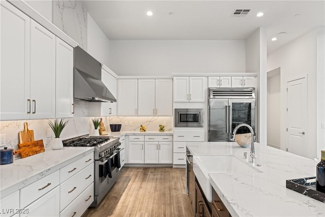 kitchen with white cabinetry, built in appliances, light stone counters, wall chimney range hood, and light hardwood / wood-style flooring