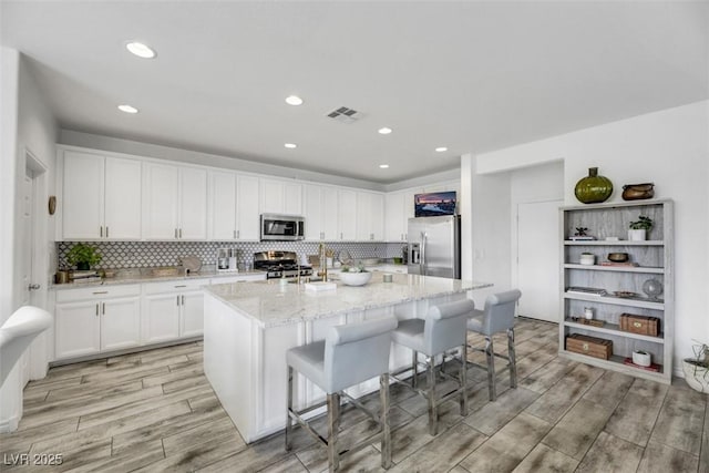 kitchen featuring white cabinetry, appliances with stainless steel finishes, a kitchen breakfast bar, light stone countertops, and a kitchen island with sink