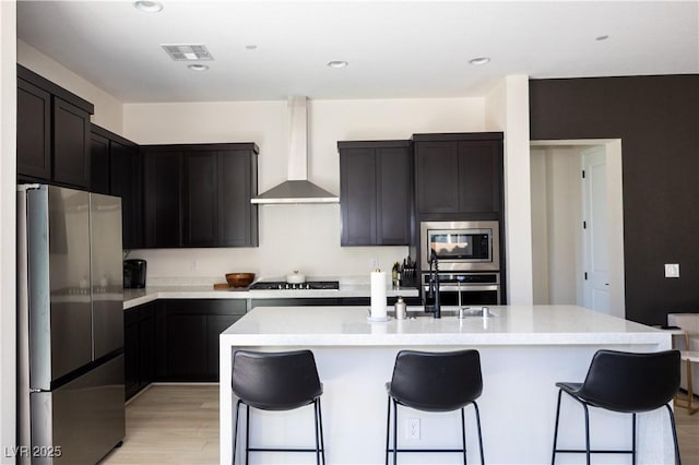 kitchen with wall chimney exhaust hood, appliances with stainless steel finishes, a kitchen bar, and an island with sink