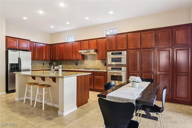 kitchen with reddish brown cabinets, under cabinet range hood, a center island with sink, appliances with stainless steel finishes, and a sink