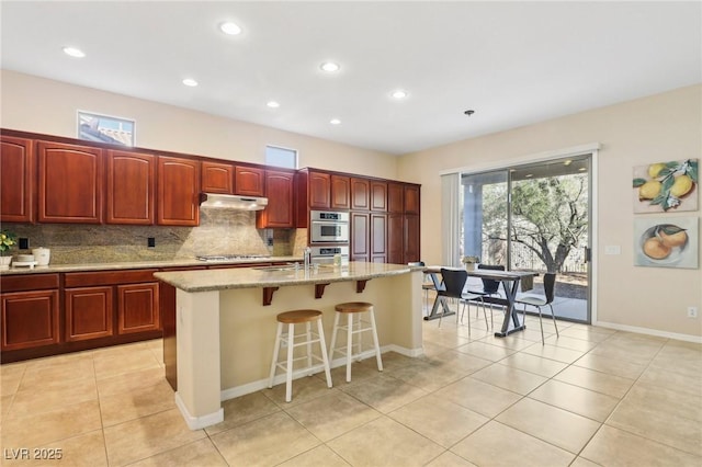 kitchen featuring stainless steel appliances, light stone counters, an island with sink, a kitchen bar, and decorative backsplash