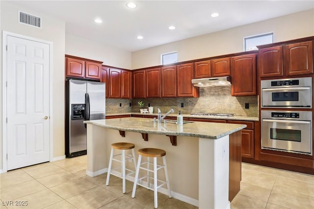 kitchen with sink, a center island with sink, light tile patterned floors, appliances with stainless steel finishes, and a kitchen breakfast bar