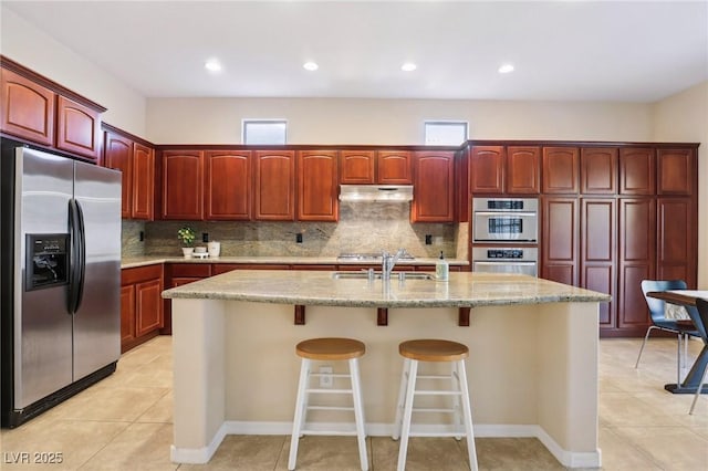 kitchen with light tile patterned flooring, sink, backsplash, stainless steel appliances, and a center island with sink