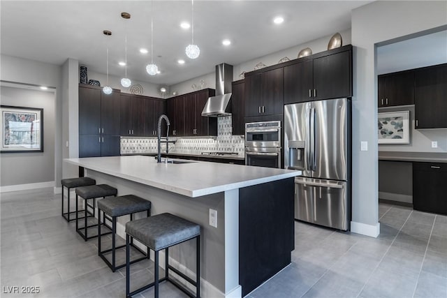 kitchen with pendant lighting, wall chimney range hood, stainless steel appliances, dark brown cabinetry, and an island with sink