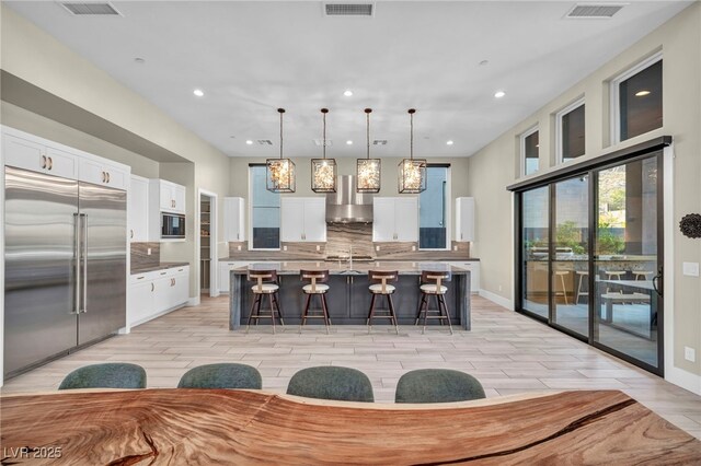 kitchen with built in appliances, hanging light fixtures, a center island with sink, and white cabinets