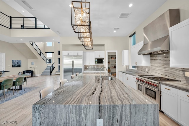 kitchen with sink, light stone counters, built in appliances, wall chimney range hood, and white cabinets