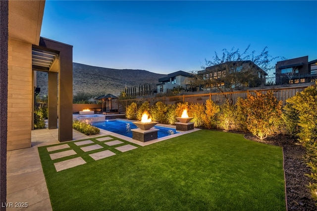 pool at dusk featuring a patio, a mountain view, a lawn, and a fire pit