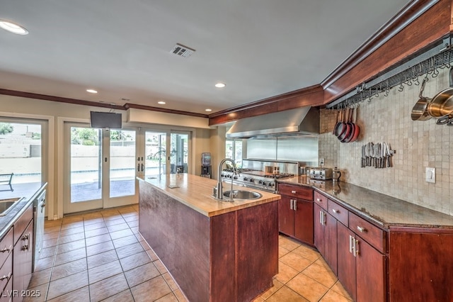 kitchen with a sink, visible vents, reddish brown cabinets, wall chimney exhaust hood, and an island with sink