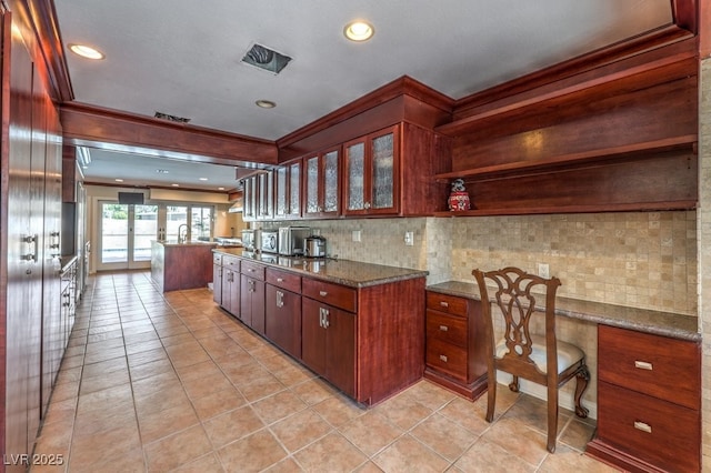 kitchen featuring visible vents, dark stone counters, built in study area, glass insert cabinets, and backsplash