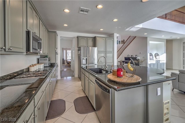 kitchen with sink, light tile patterned floors, dark stone countertops, stainless steel appliances, and an island with sink