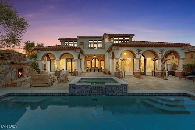 back house at dusk with a balcony, a swimming pool with hot tub, an outdoor living space with a fireplace, and a patio area